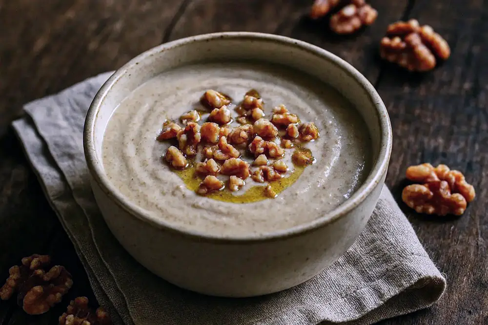 A white ceramic bowl of pale, creamy walnut tarator garnished with crushed walnuts and a drizzle of sunflower oil, on a linen cloth